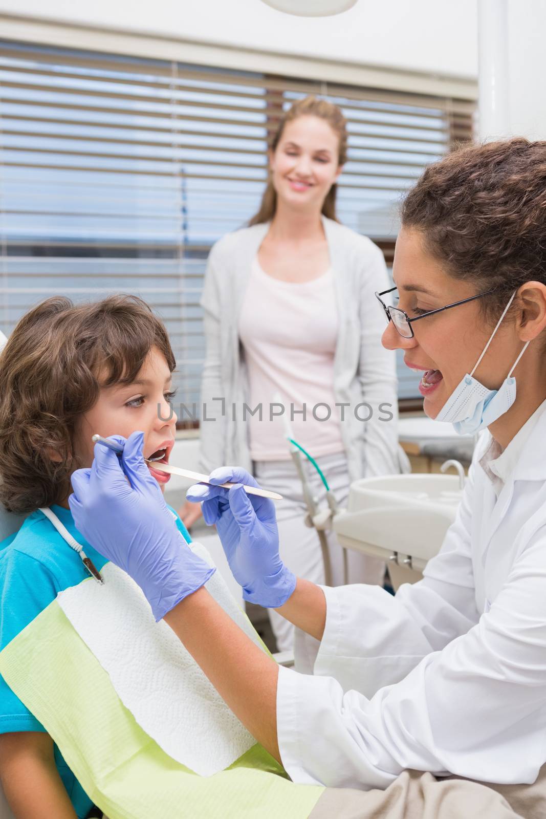 Pediatric dentist examining a little boys teeth with his mother watching at the dental clinic