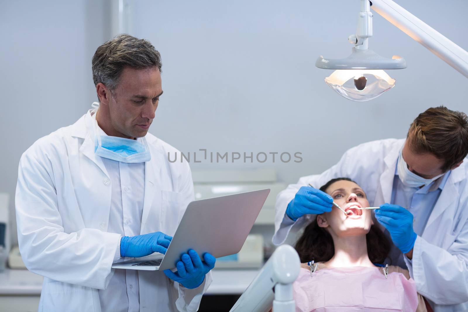 Dentist using laptop while colleague examining a female patient at dental clinic