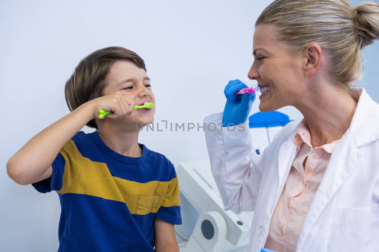 Happy dentist and boy brushing teeth against wall at clinic 