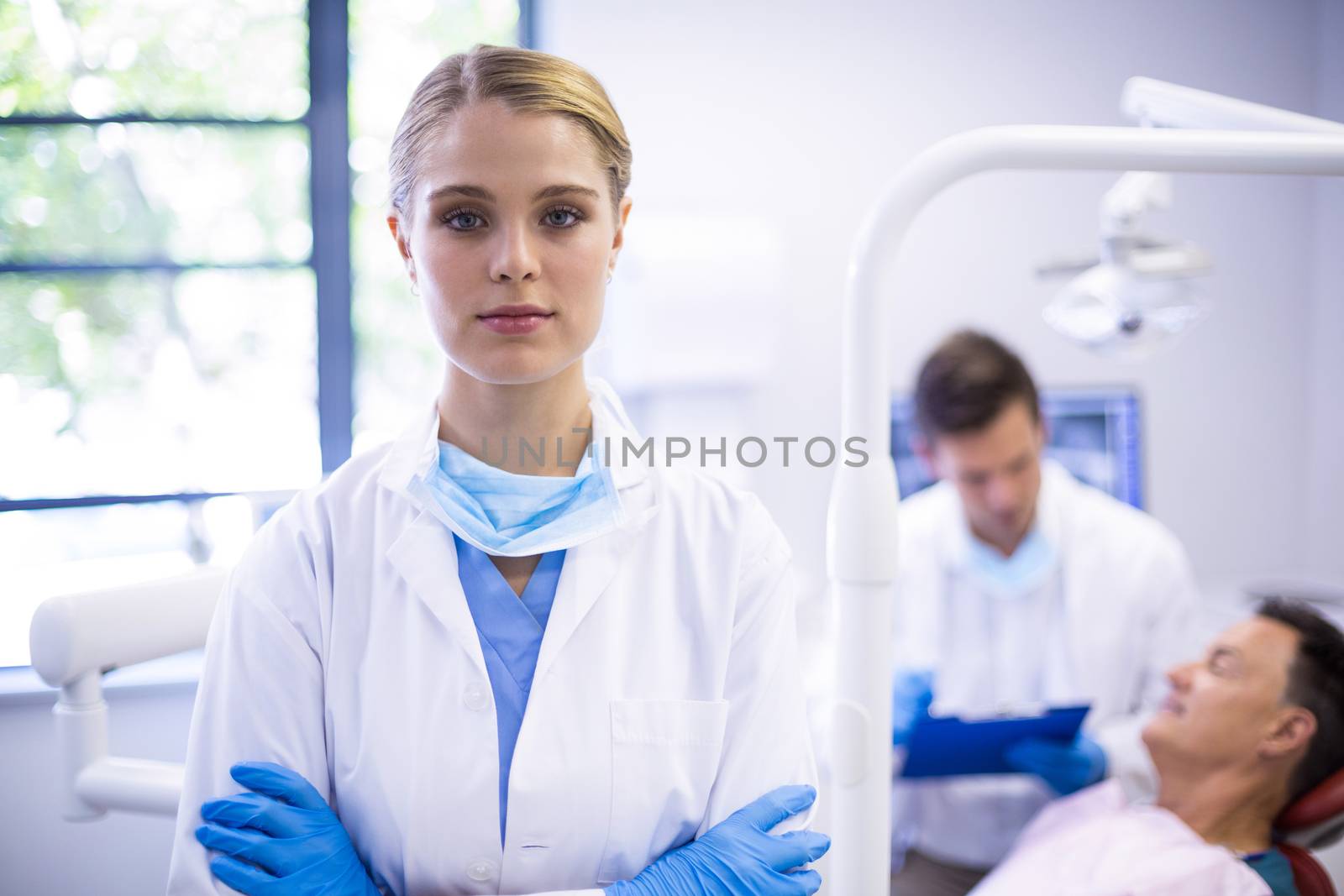 Portrait of dentist standing with arms crossed while her colleague examining patient in background
