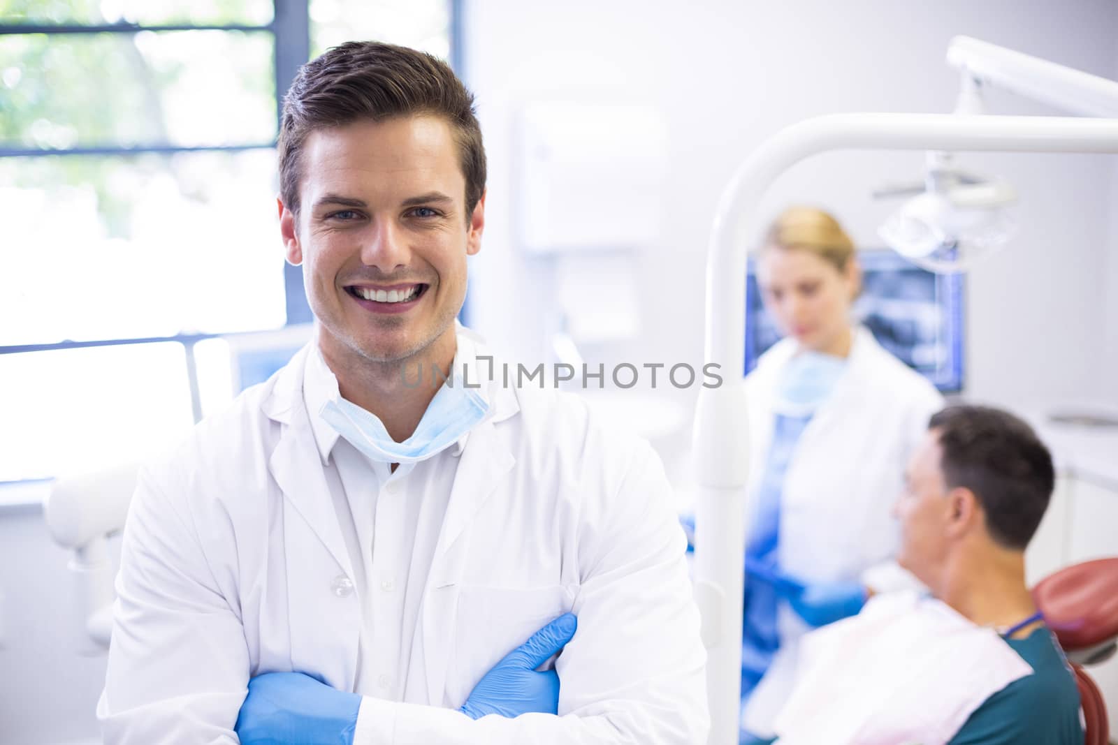 Portrait of dentist standing with arms crossed while his colleague examining patient in background