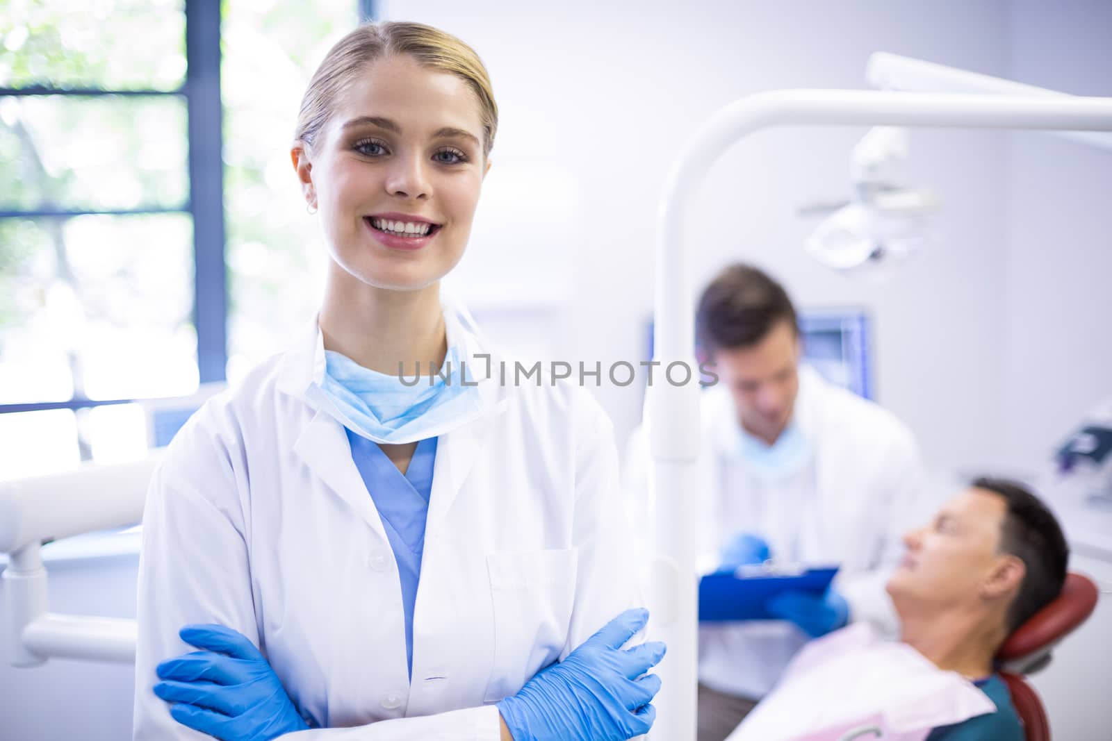 Portrait of smiling dentist standing  with arms crossed while her colleague examining patient in background
