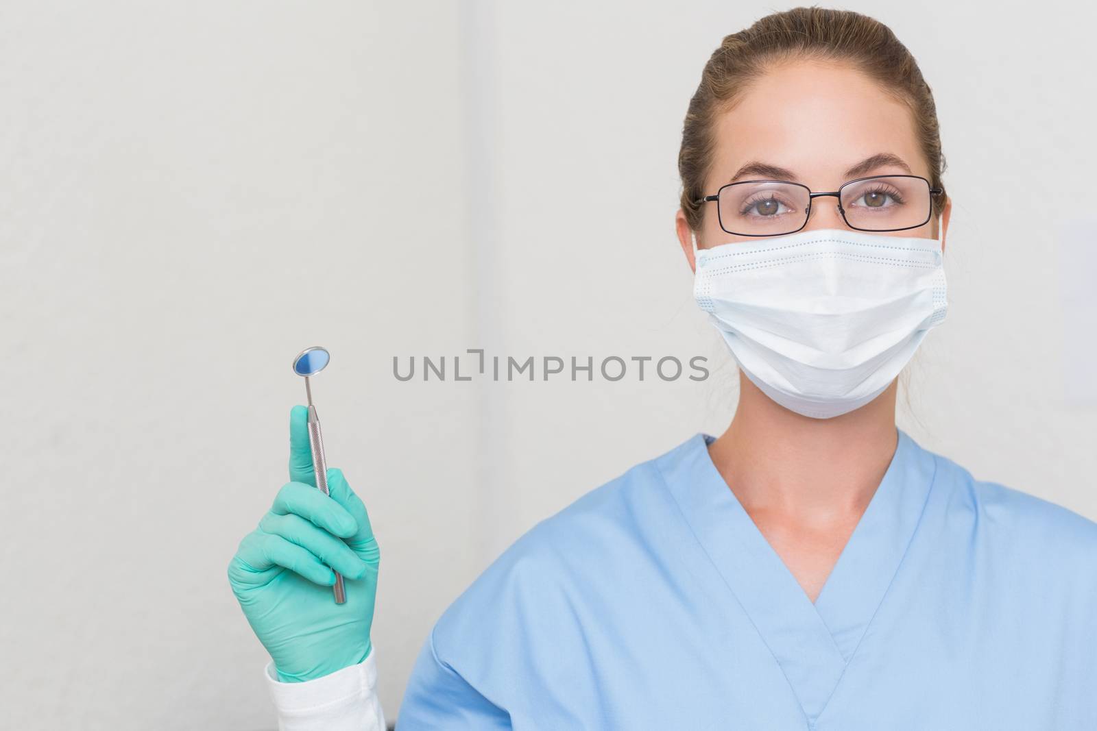 Dentist in blue scrubs holding angled mirror looking at camera at the dental clinic
