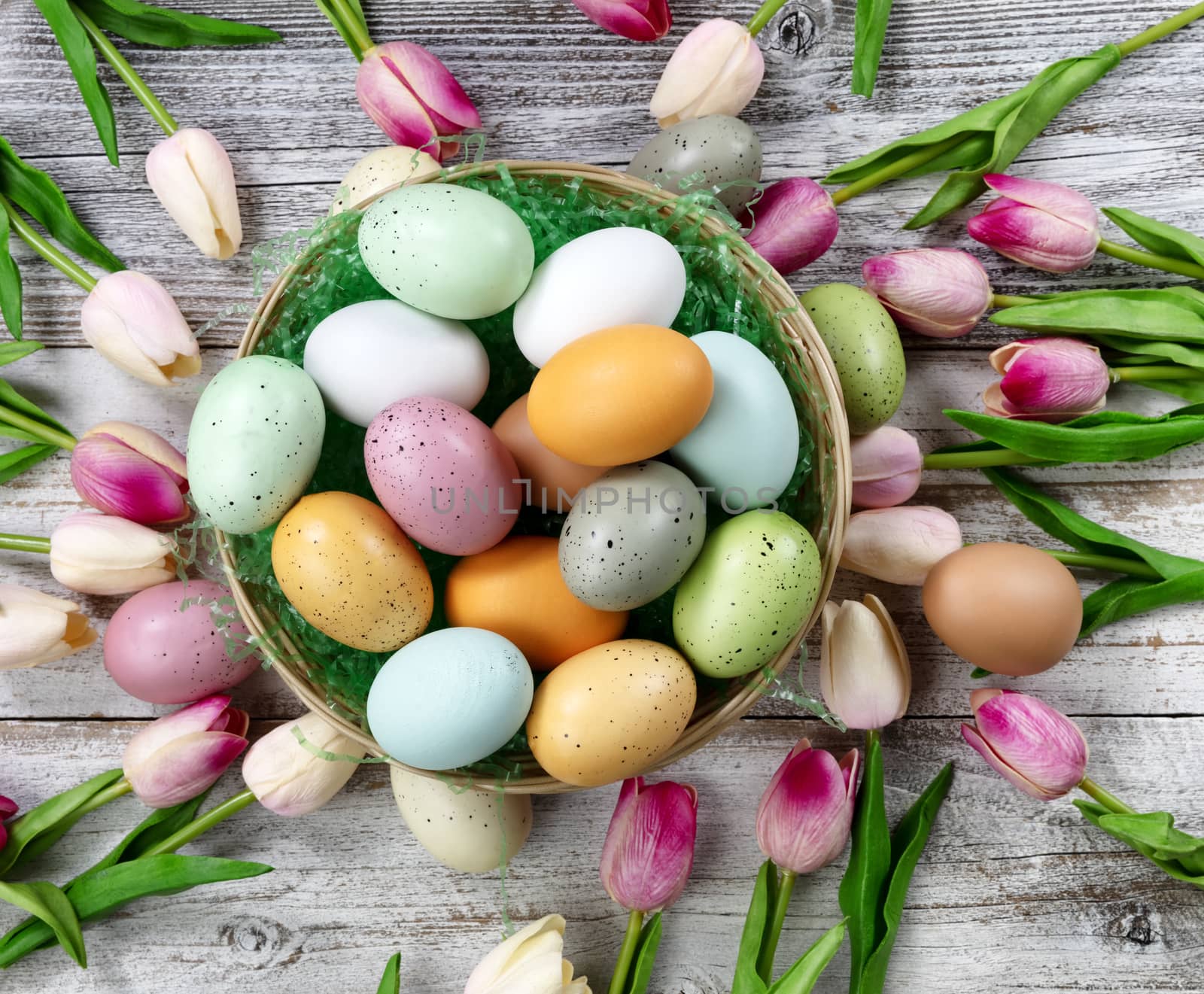 Happy Easter concept with basket filled of colorful eggs and spring flowers on white rustic wood. Overhead view layout 
