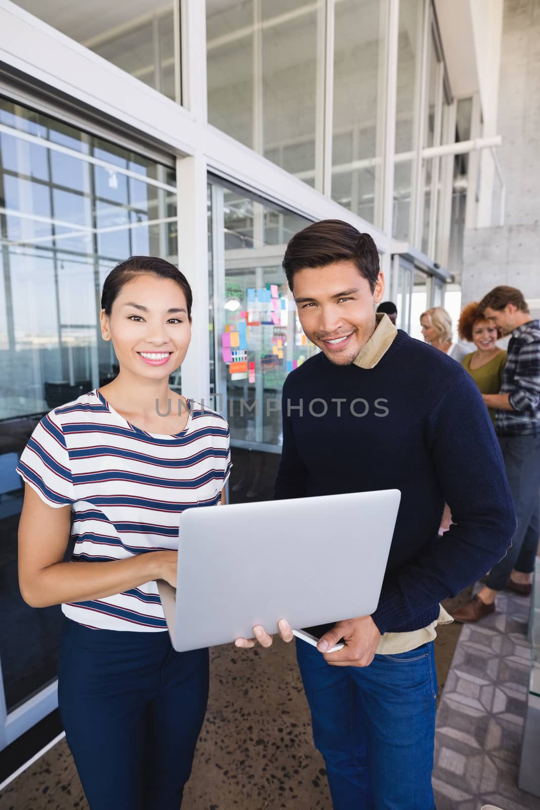 Portrait of smiling business people with laptop computer standing by glass wall in office