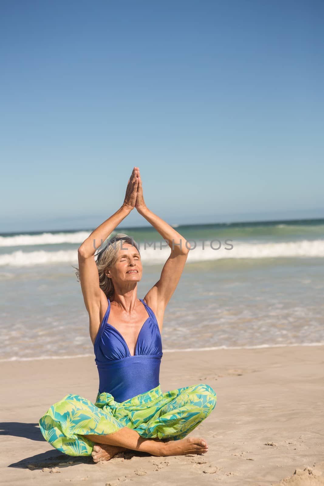 Senior woman practising yoga while sitting against sea at beach