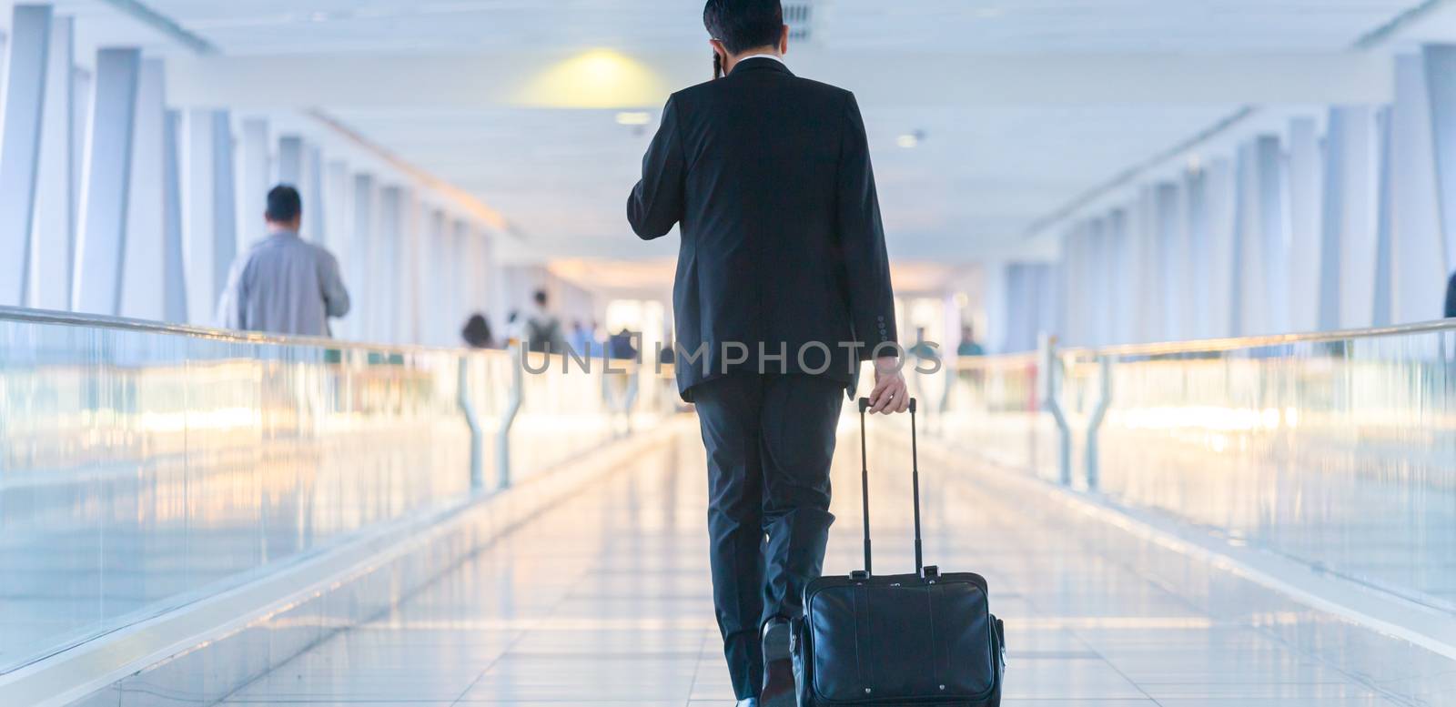 Rear view of unrecognizable formaly dressed businessman walking and wheeling a trolley suitcase at the lobby, talking on a mobile phone. Business travel concept.