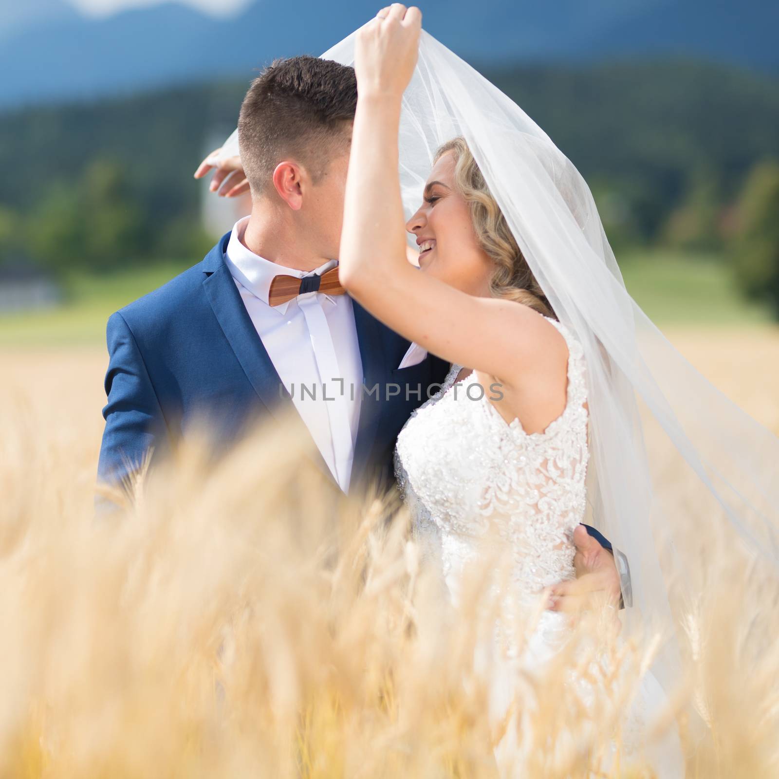 Groom hugs bride tenderly while wind blows her veil in wheat field somewhere in Slovenian countryside. Caucasian happy romantic young couple celebrating their marriage.