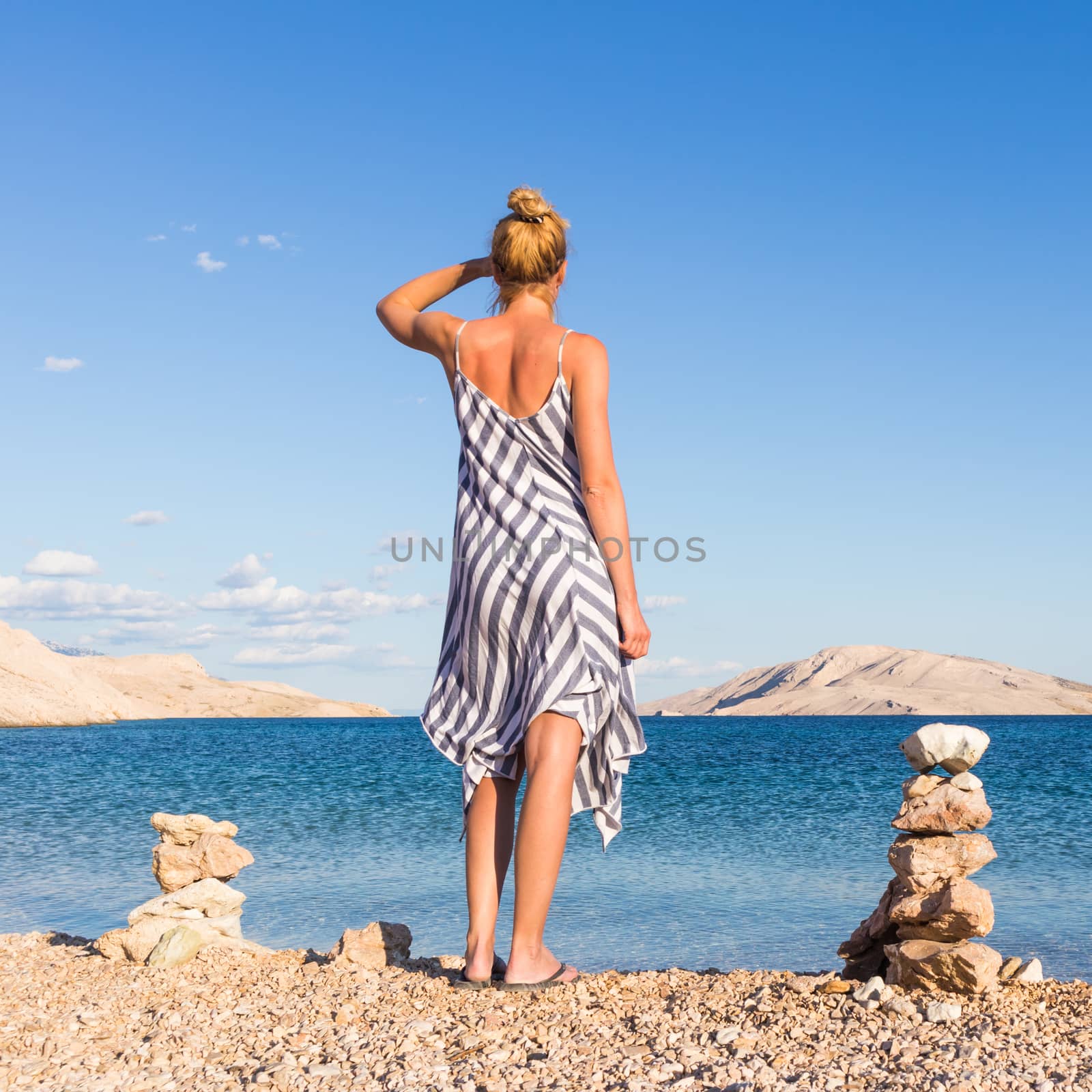 Happy carefree woman wearing beautiful striped summer dress enjoying late afternoon walk on white pabbled beach on Pag island, Croatia.