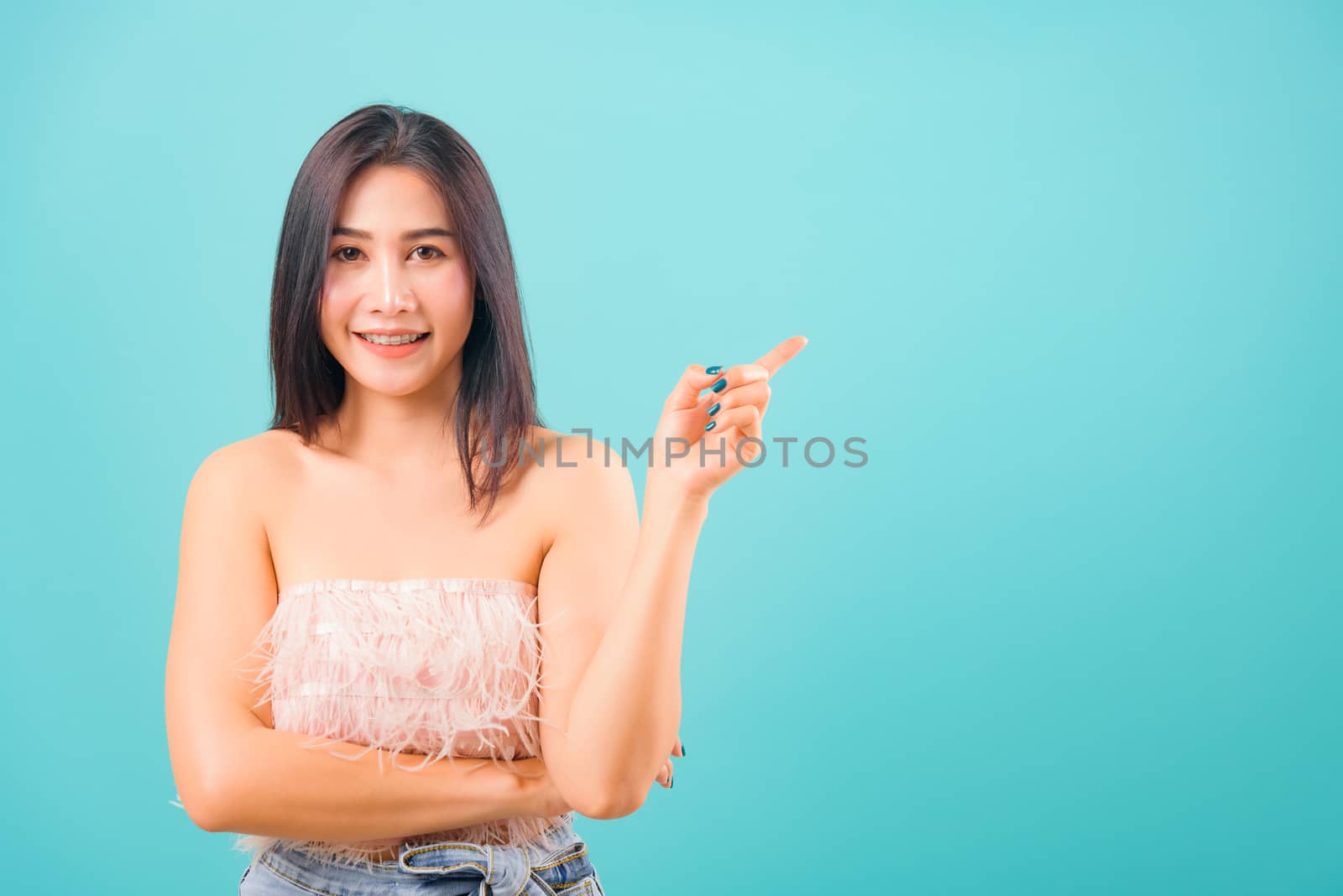 Smiling face Asian beautiful woman her point finger beside and looking to camera on blue background, with copy space for text