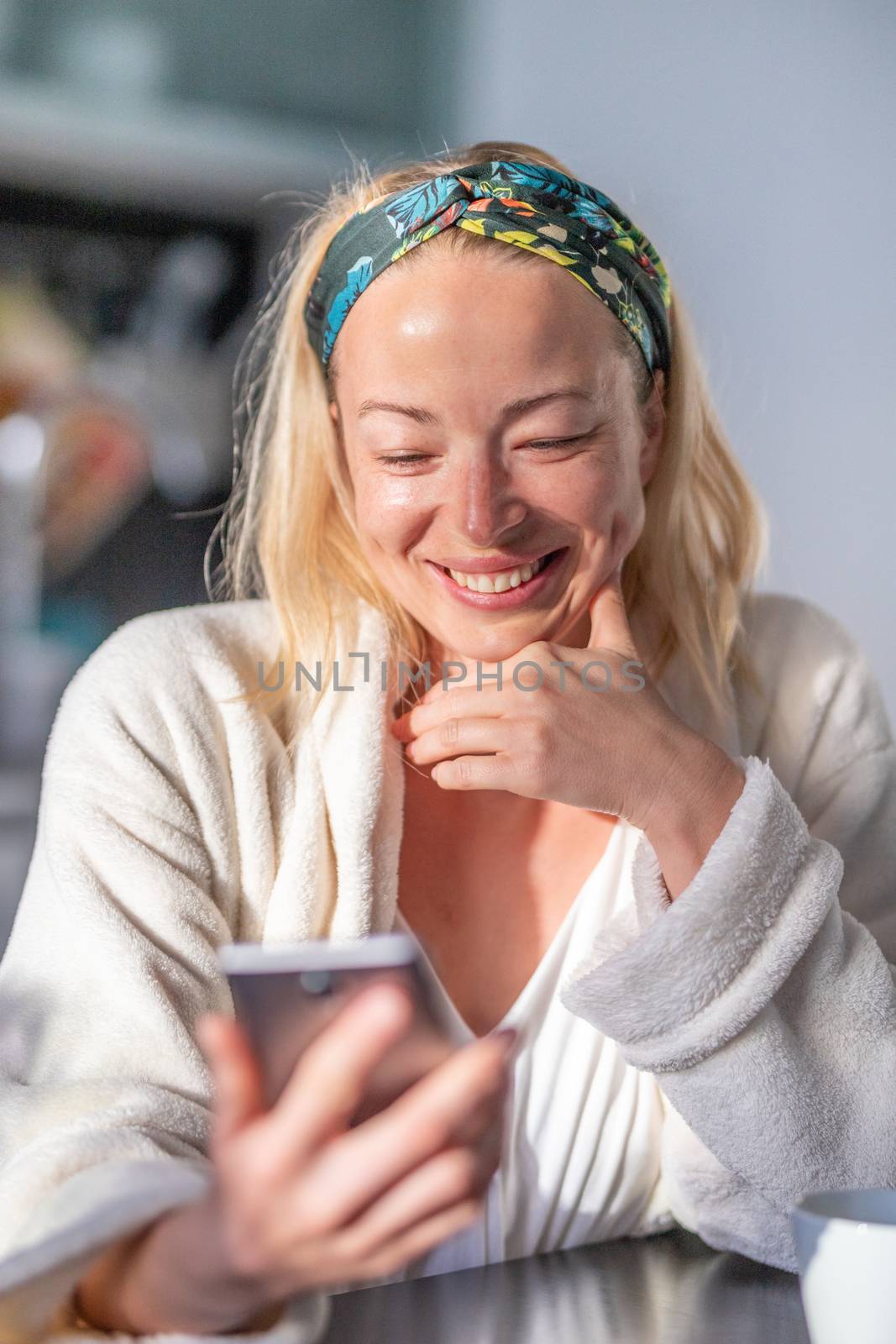 Beautiful caucasian woman at home, feeling comfortable wearing white bathrobe, taking some time to herself, drinking morning coffee and reading news on mobile phone device in the morning.