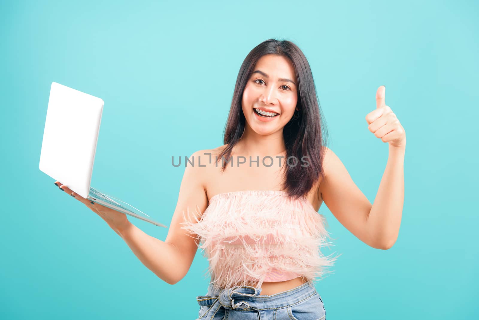 Smiling face portrait asian beautiful woman holding laptop computer and giving thumb up on blue background, with copy space for text