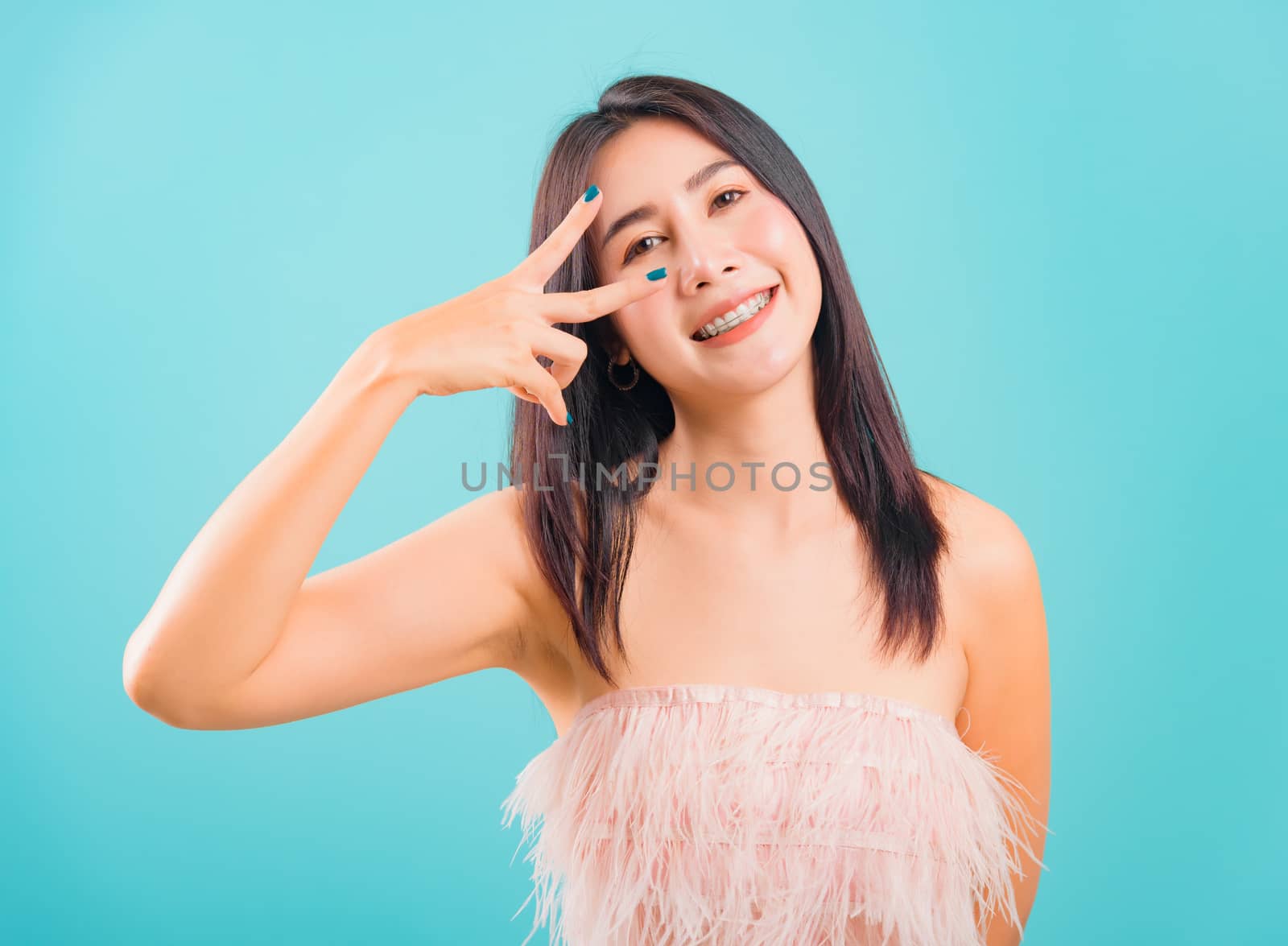 Portrait asian beautiful woman smiling showing peace gesture hand v-sign near eye good mood and her looking to camera on blue background