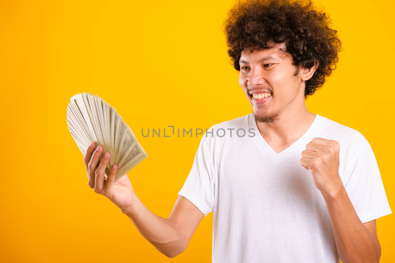 Asian handsome man with curly hair holding fans of money dollar bills isolate on yellow background with copy space for text
