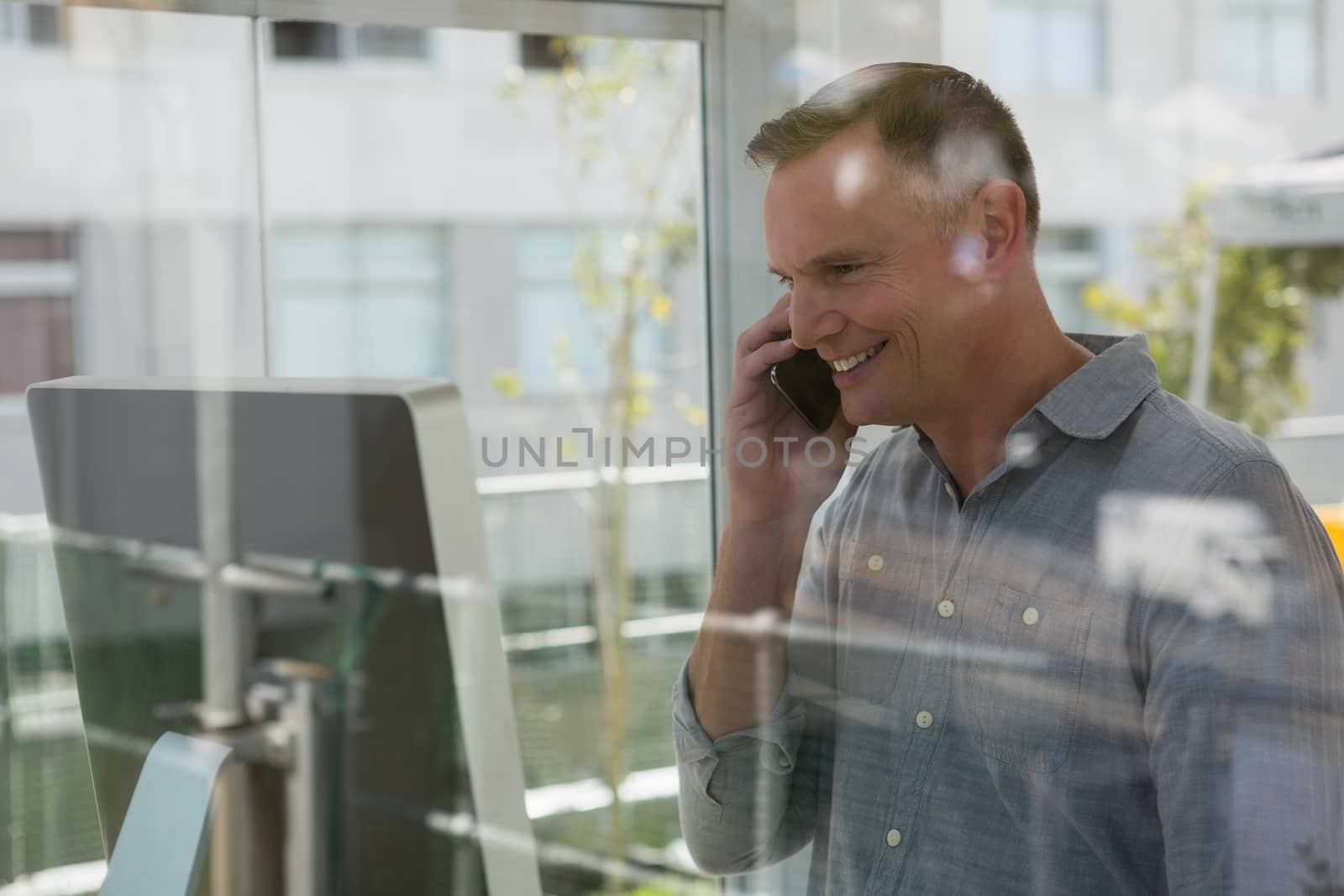 Smiling fashion designer talking on mobile phone by computer seen through glass