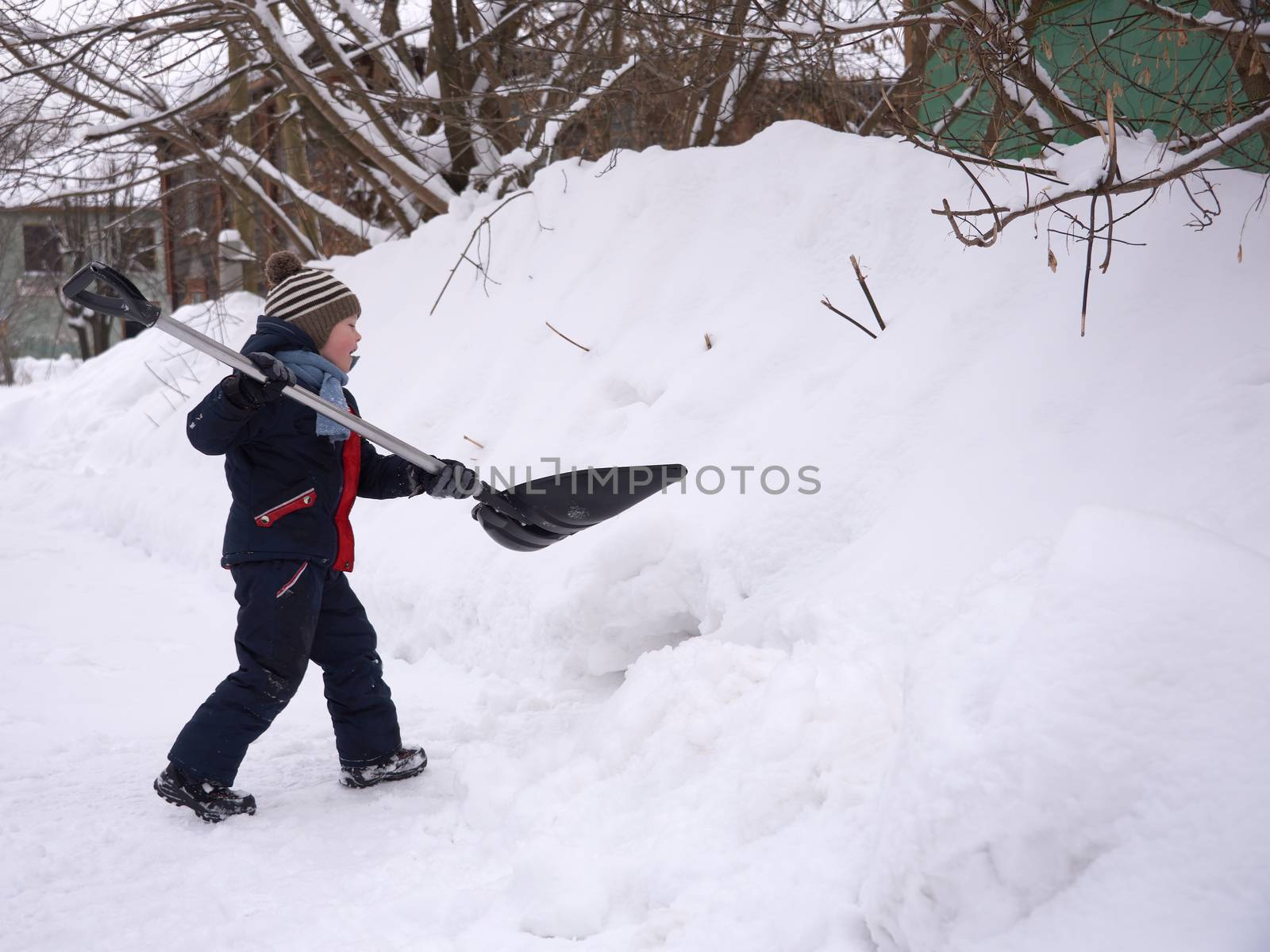 A boy in winter clothes plays with a big shovel in the snow in winter. by vladali