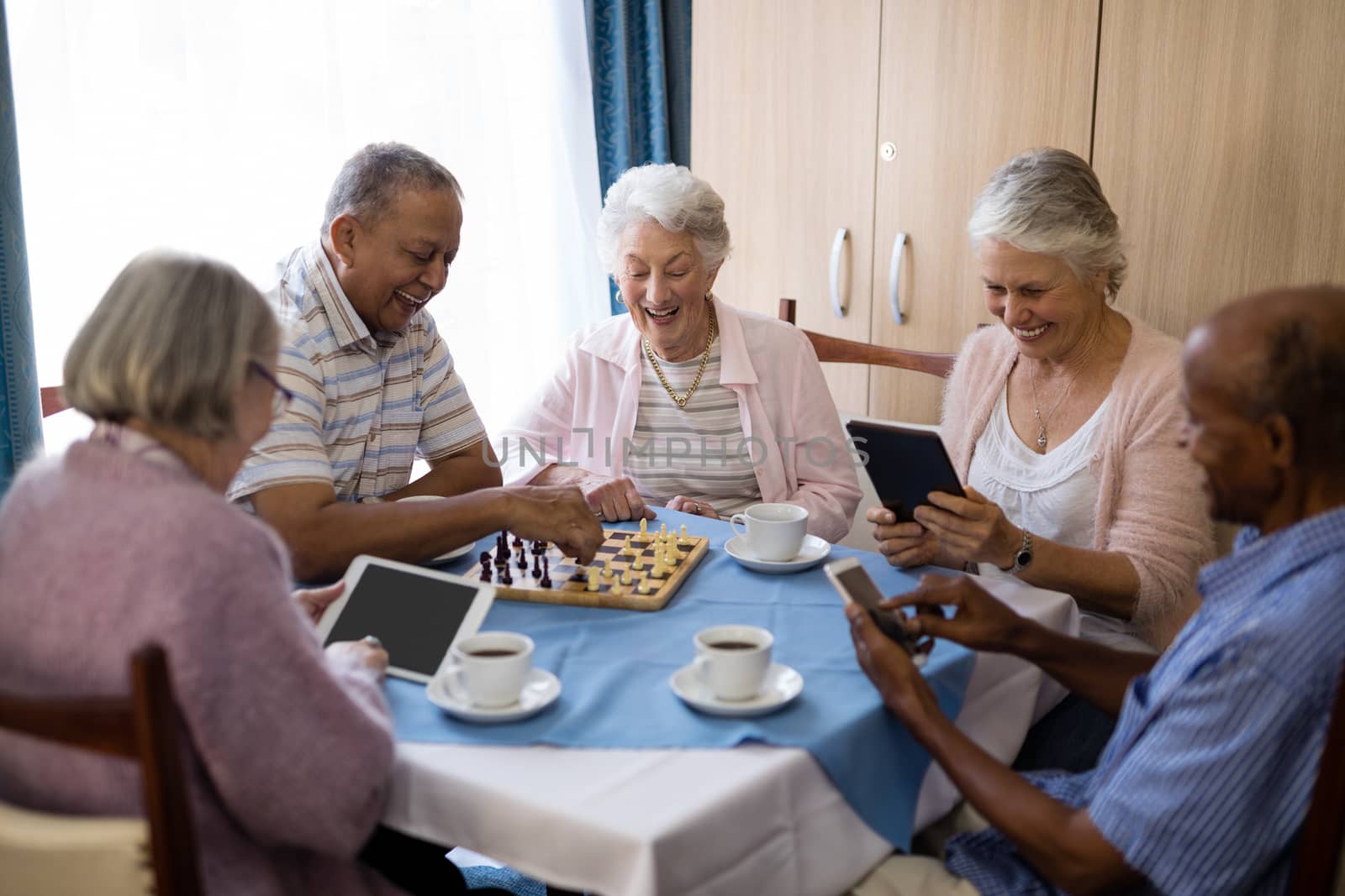 Senior friends playing chess and using technology while having coffee at table in nursing home