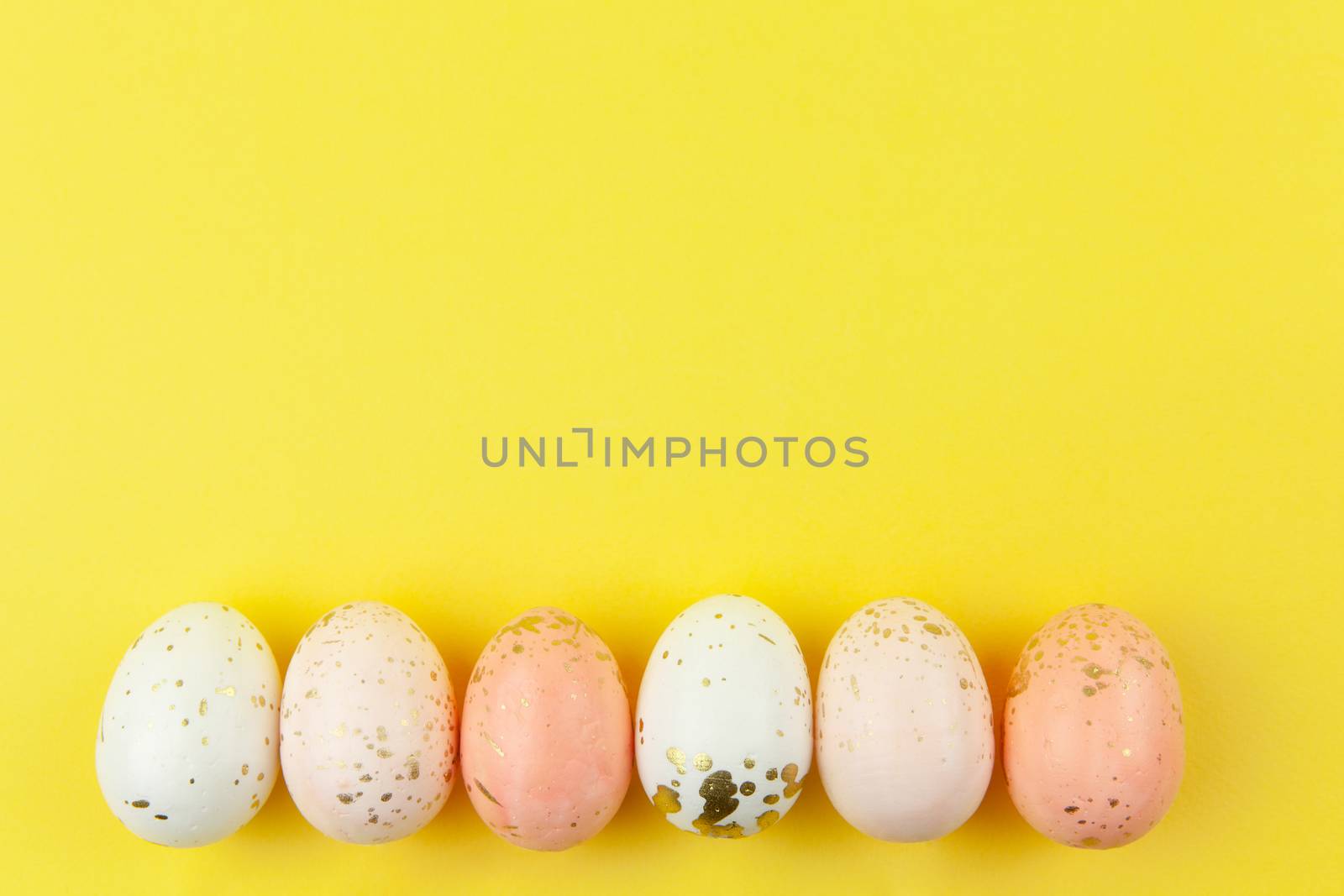 Creatively painted eggs in pastel colors decorated with gold leaf are arranged in row along bottom edge on yellow background, copy space. Happy Easter DIY concept. Flat lay. Horizontal.