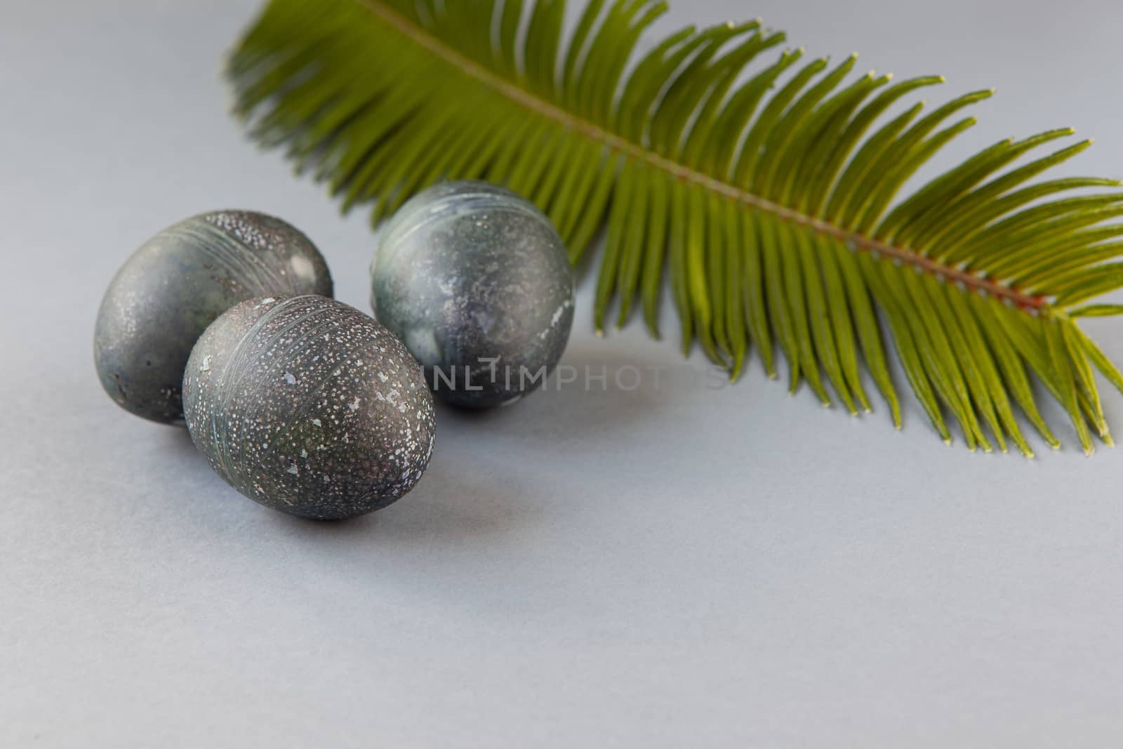 Creatively painted chicken Easter eggs with natural hibiscus dye, look like sea stones, date palm leaf on gray background, copy space. Happy creative Easter concept. Soft slective focus. Side view.