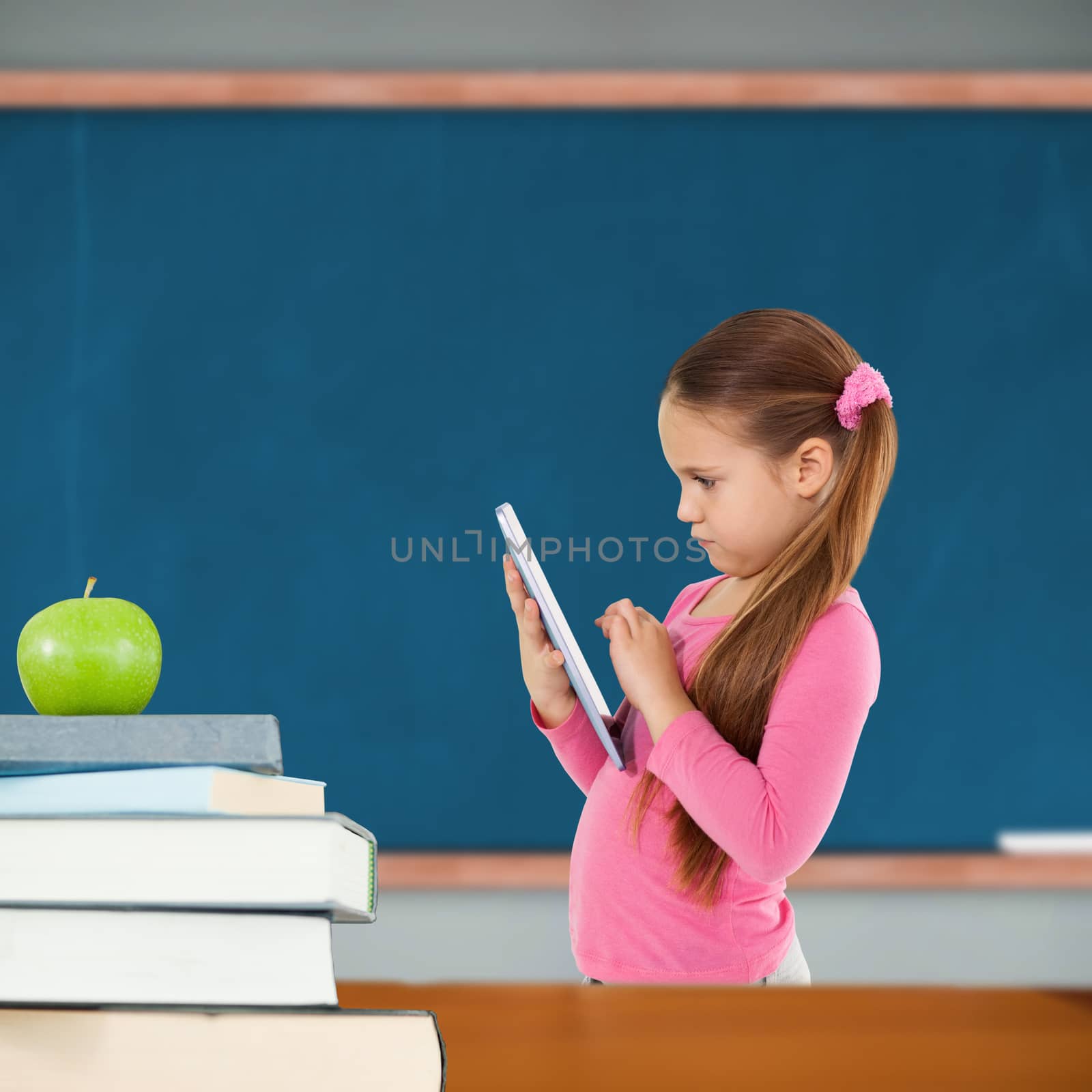 Cute girl using tablet against green apple on pile of books in classroom