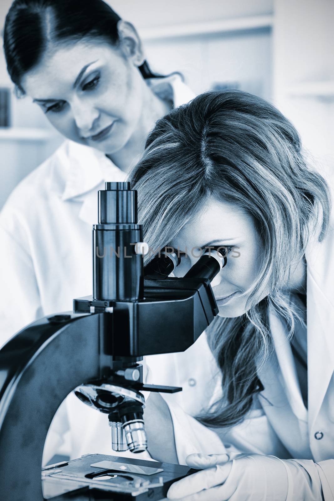 Young female scientist looking through a microscope with her assistant in a lab