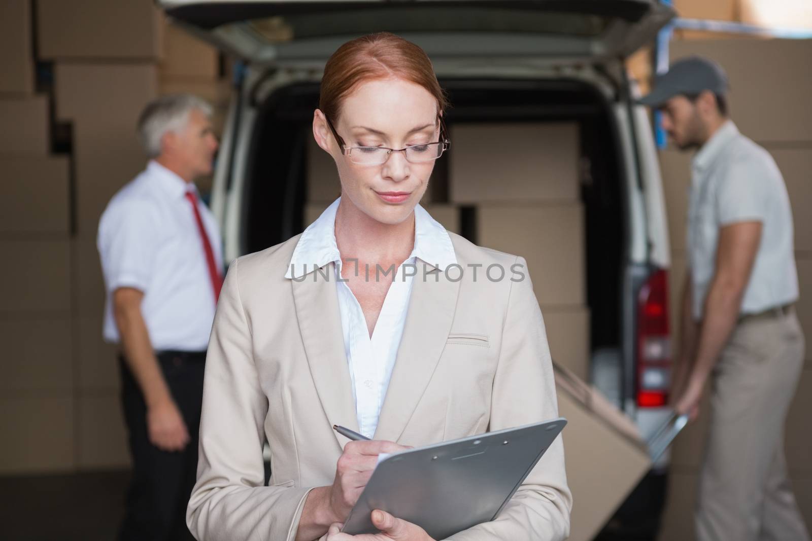 Warehouse manager writing in clipboard with delivery in background by Wavebreakmedia