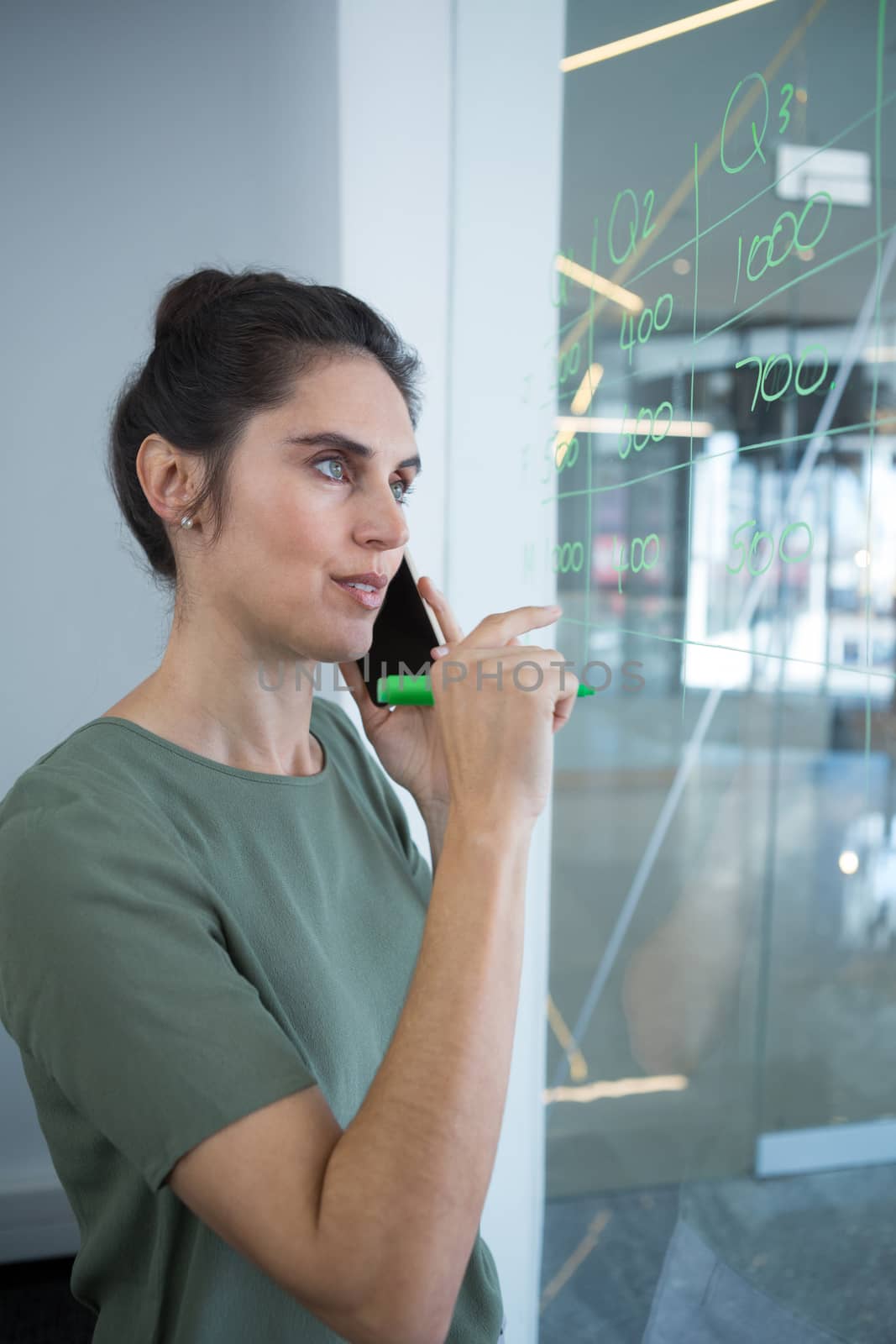 Female executive talking on mobile phone while writing on glass in office