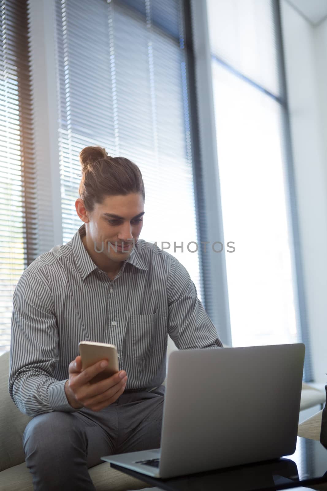Male executive holding mobile phone while using laptop in office
