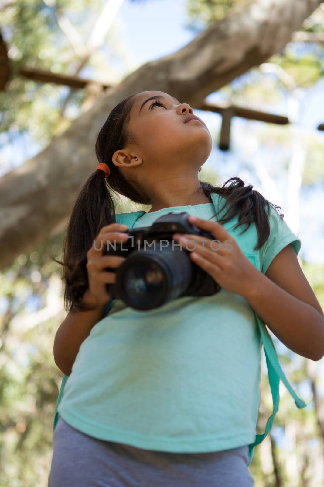 Little girl with backpack holding dslr camera in her hand looking up in the sky on a sunny day in the forest