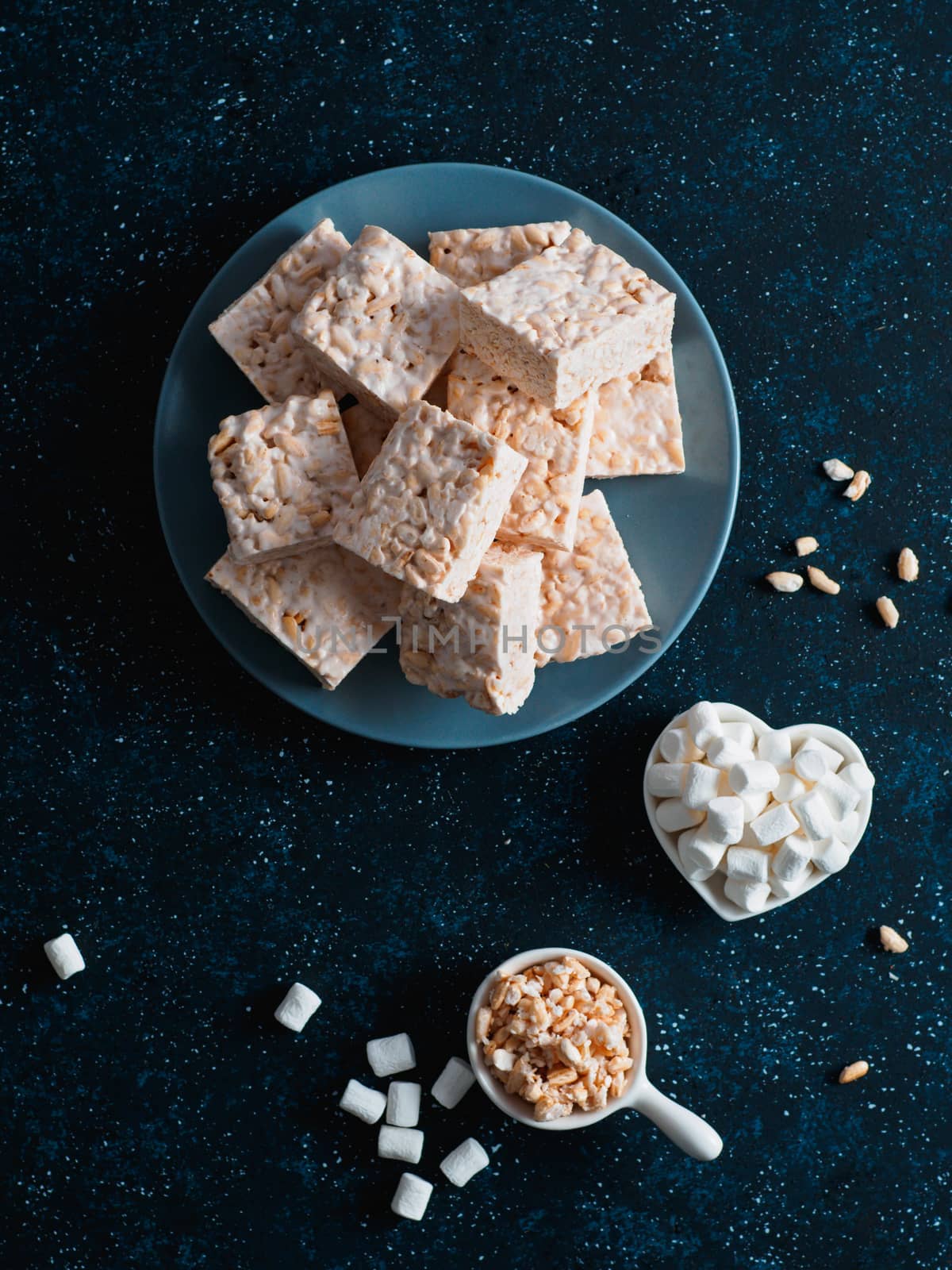 Homemade square bars of Marshmallow and crispy rice and ingredients on dark blue background. American dessert with marshmallow and crispy rice. Top view. Copy space