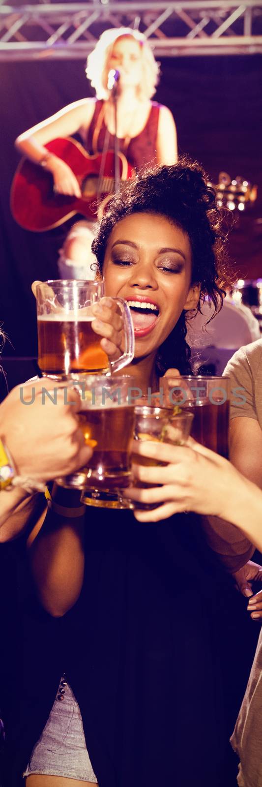 Smiling friends toasting beer glasses with performer singing in background at nightclub