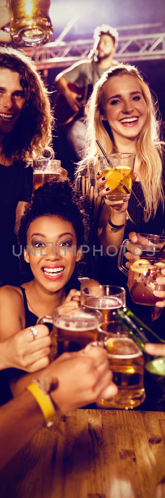 Portrait of cheerful friends toasting drinks at table with performer singing on stage in nightclub