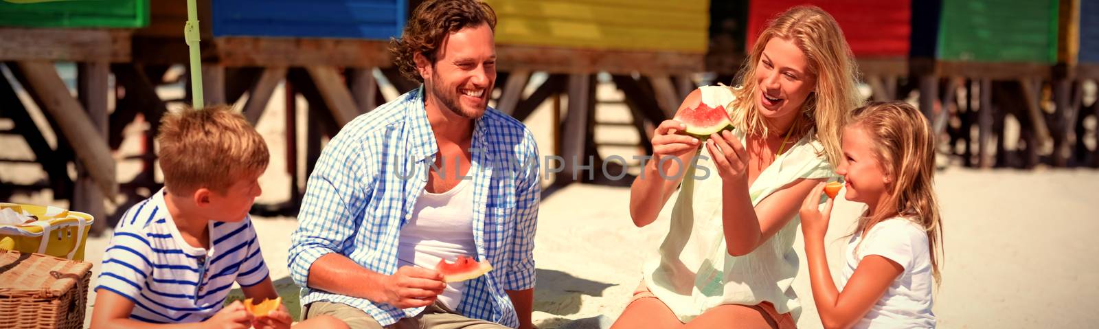 Happy family eating watermelon while sitting together on blanket at beach during sunny day