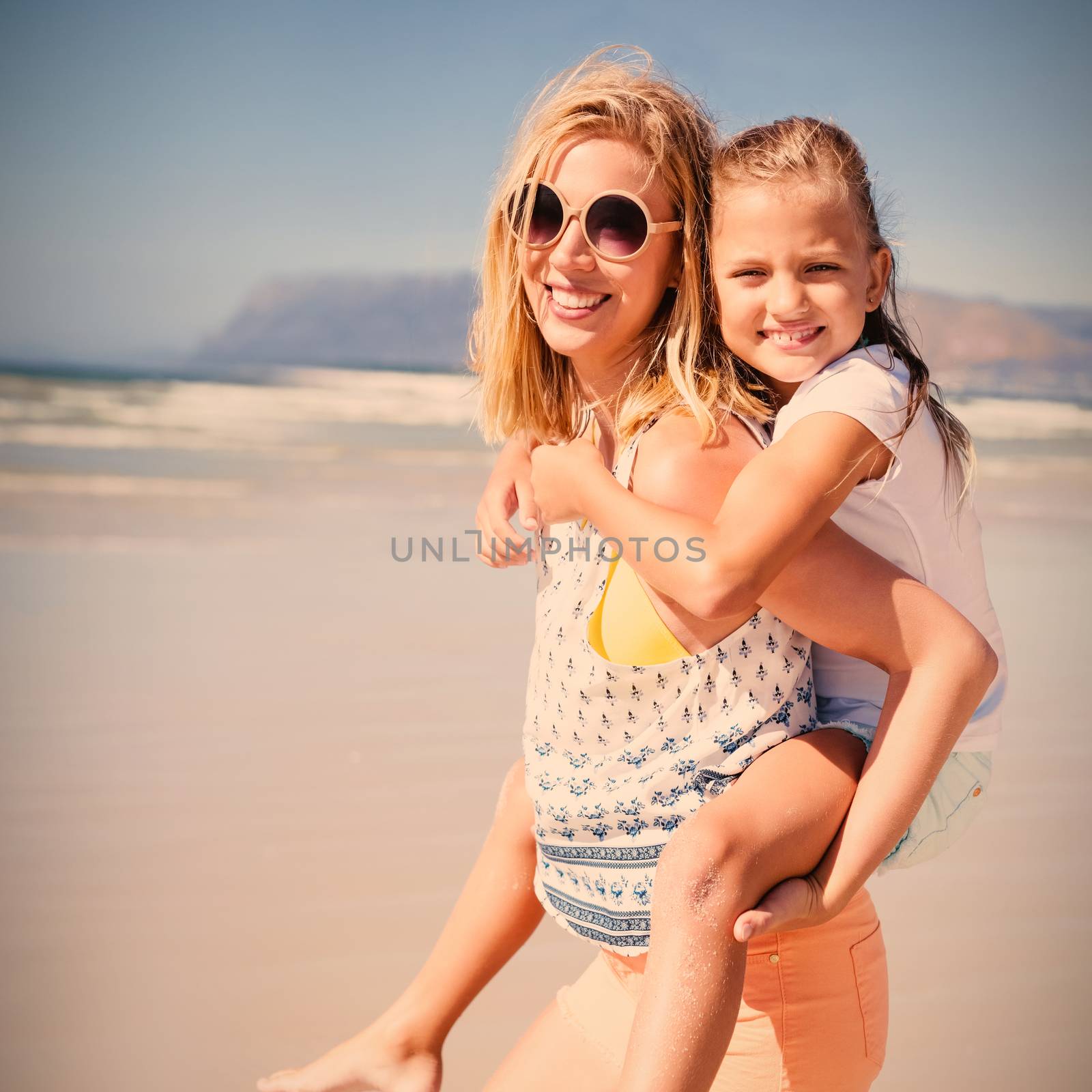 Portrait of happy mother piggybacking her daughter at beach during sunny day