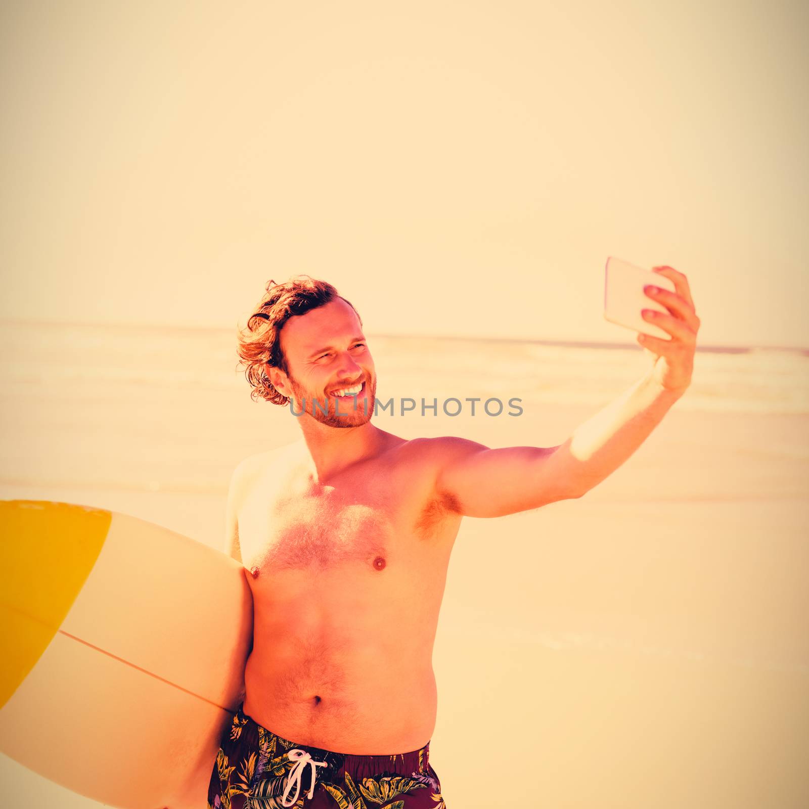 Smiling shirtless man taking selfie with surfboard at beach during sunny day