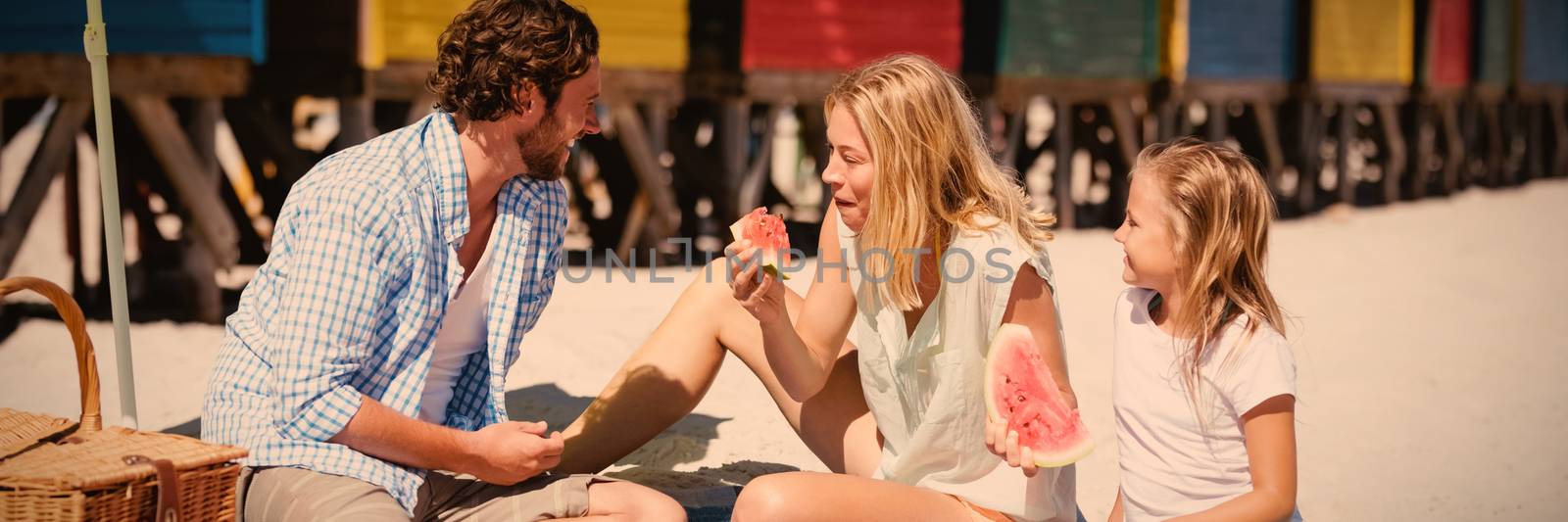 Happy family eating watermelon while sitting on blanket at beach during sunny day