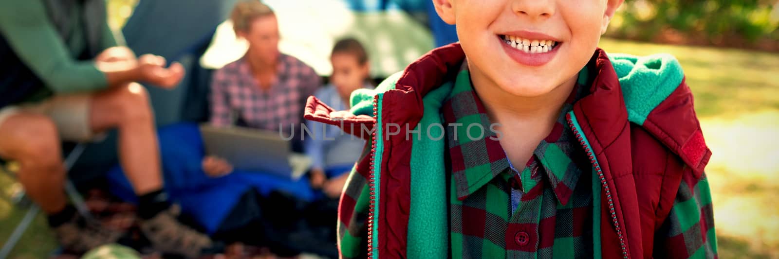 Boy smiling at camera while family sitting at tent in background on a sunny day