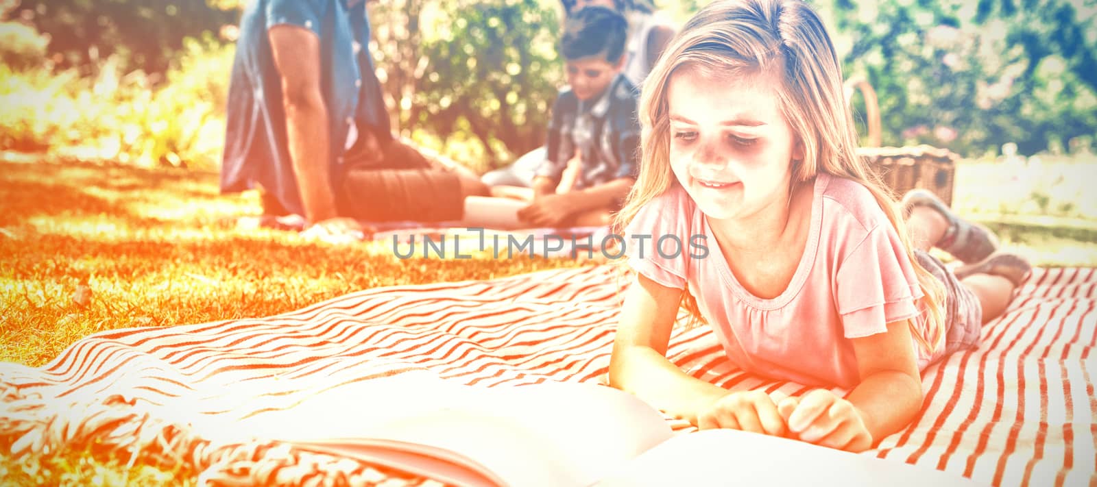 Girl lying on blanket and reading book while family sitting in background by Wavebreakmedia