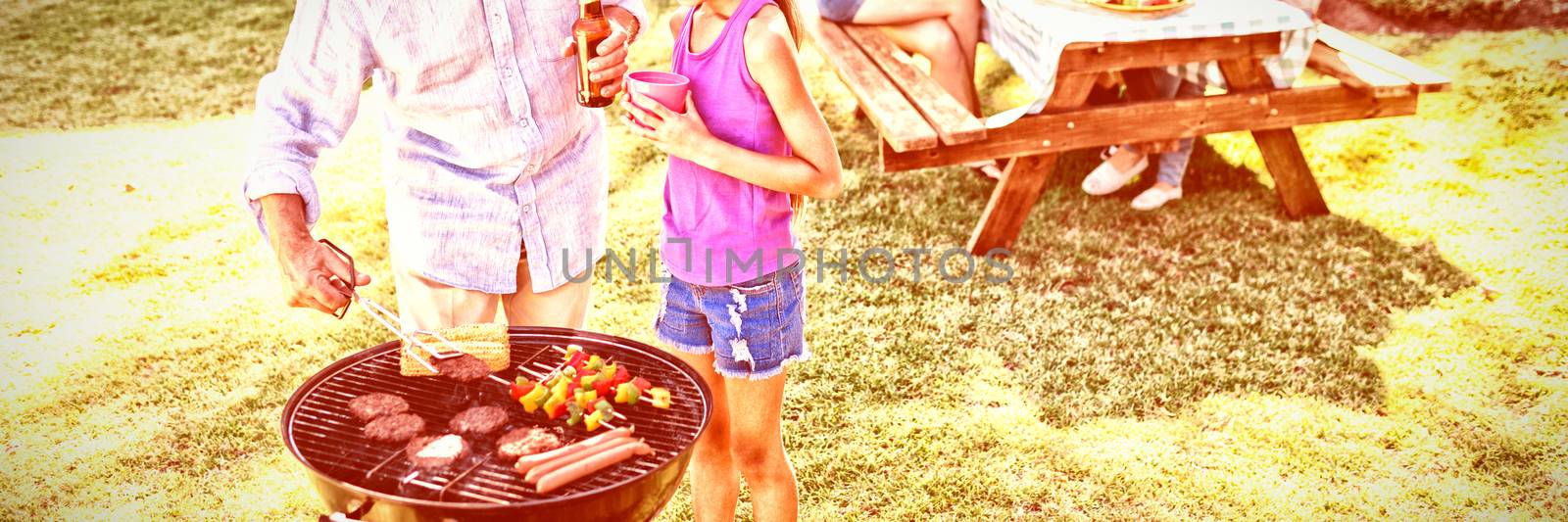 Grandfather and granddaughter preparing barbecue while family having meal by Wavebreakmedia