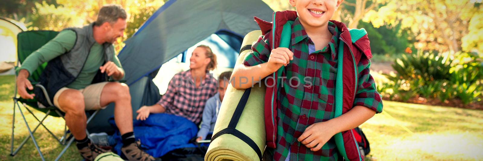 Portrait of boy holding a rolled mat while family sitting in the tent