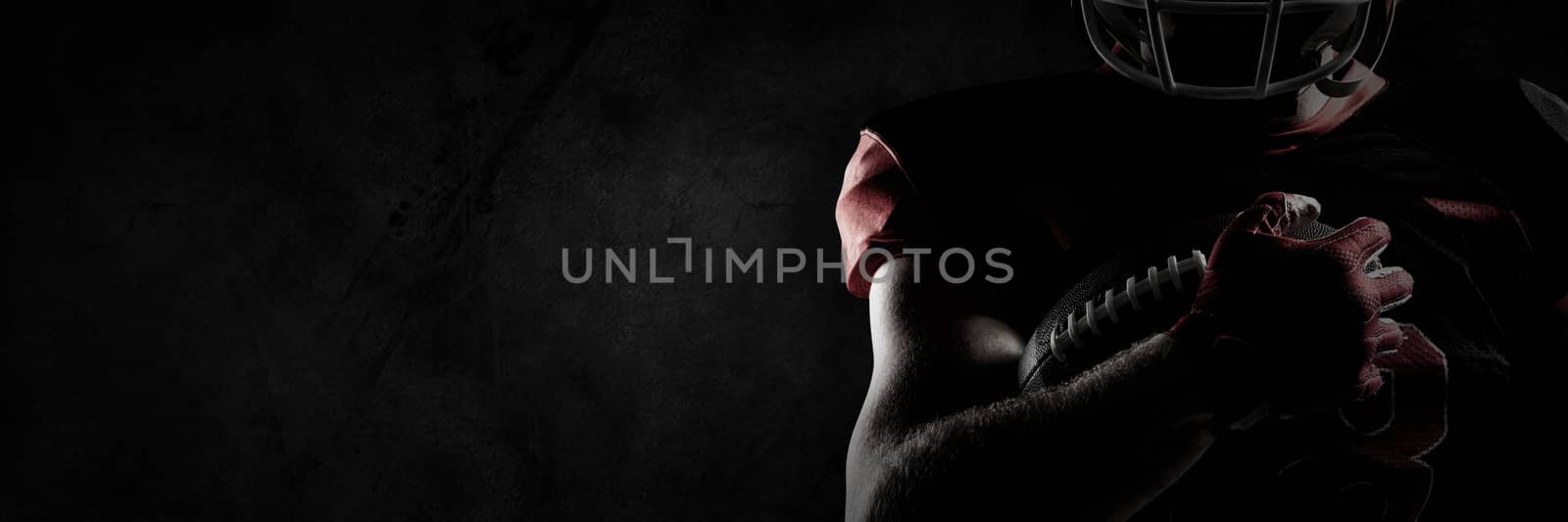 American football player in helmet holding rugby ball with hand raised against close up of grunged wall