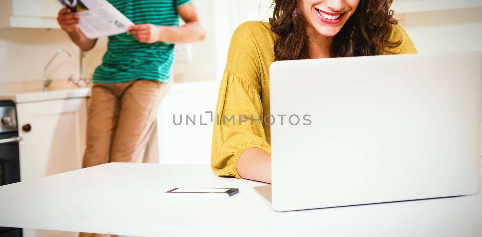 Woman using laptop while man reading newspaper in background at kitchen