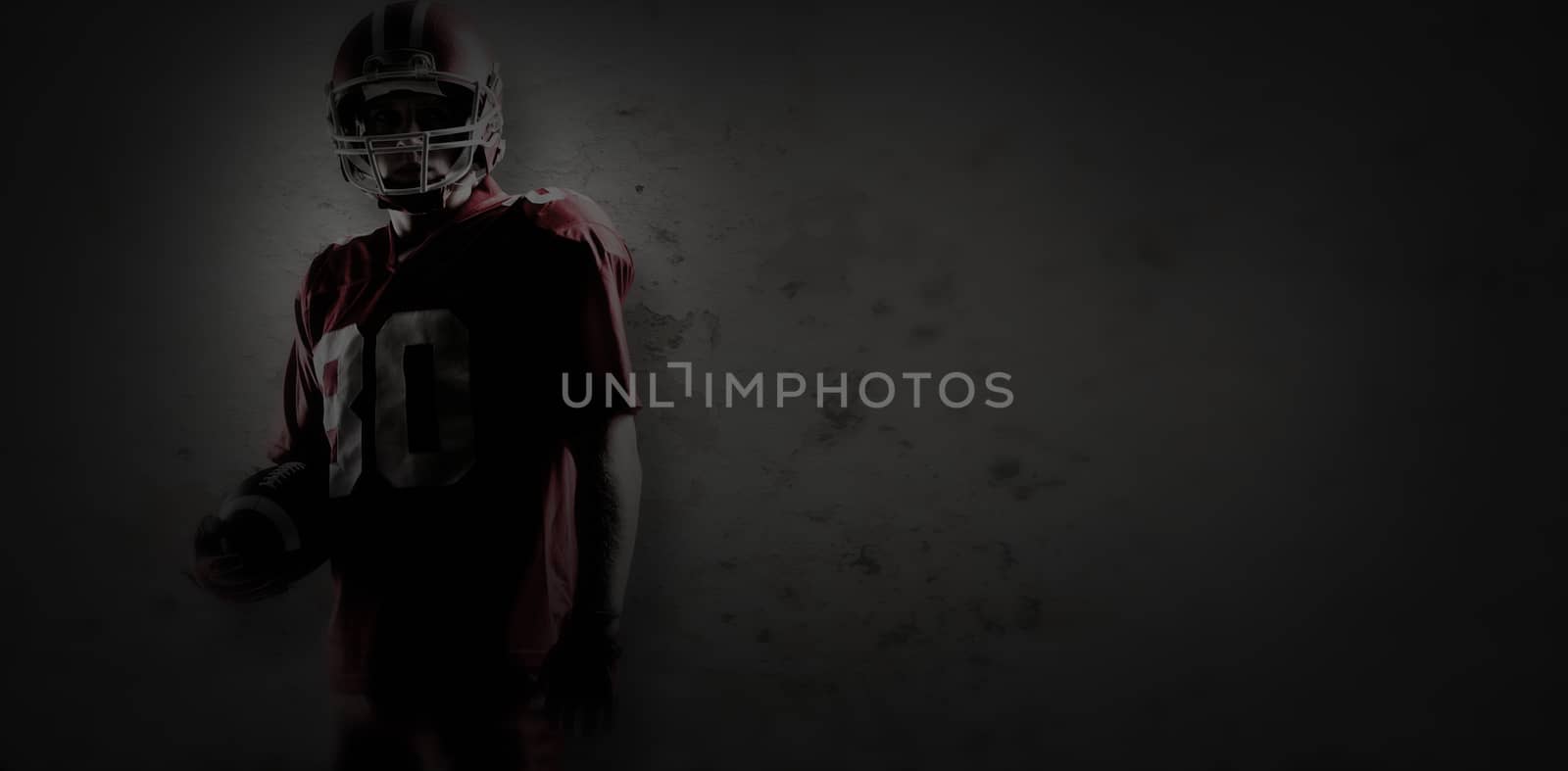 American football player in helmet holding rugby ball against old weathered wall