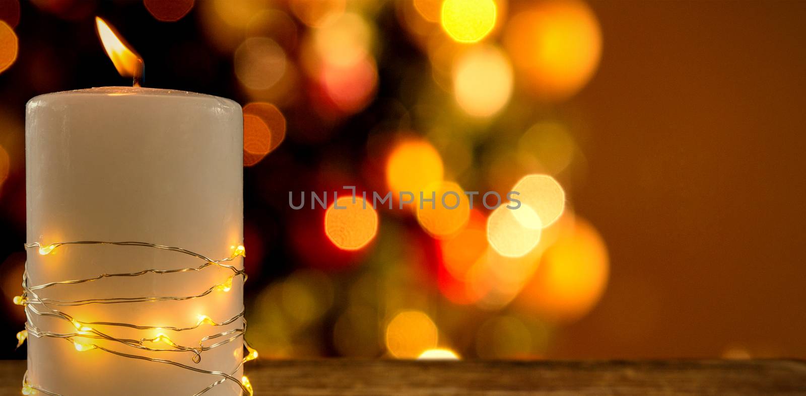 Illuminated string lights wrapped on lit candle against two red christmas balls on a white fur