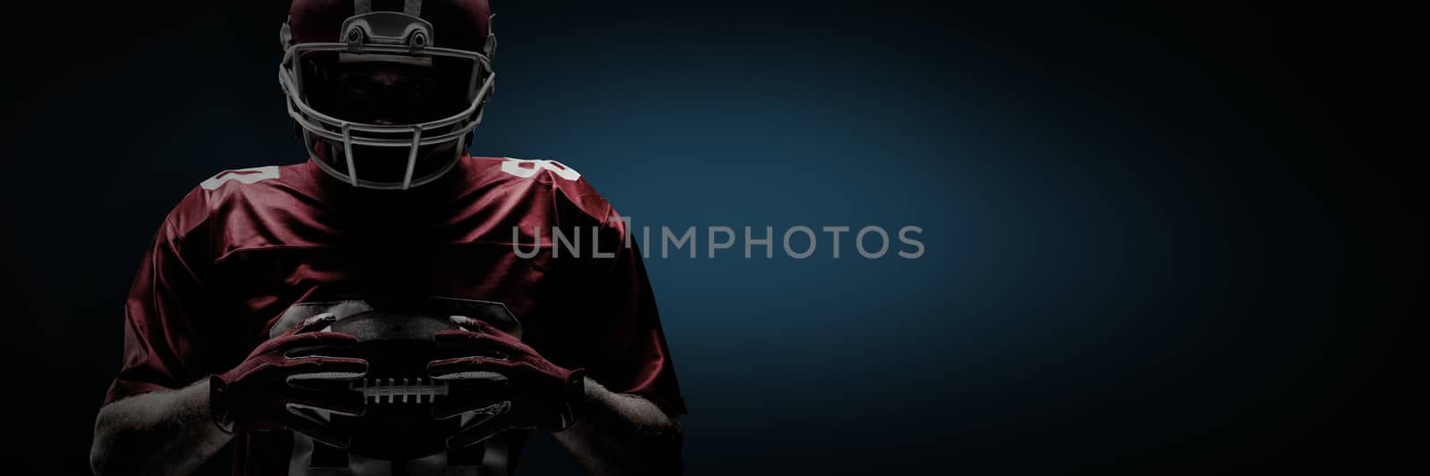 American football player standing in rugby helmet and holding rugby ball against blue background with vignette
