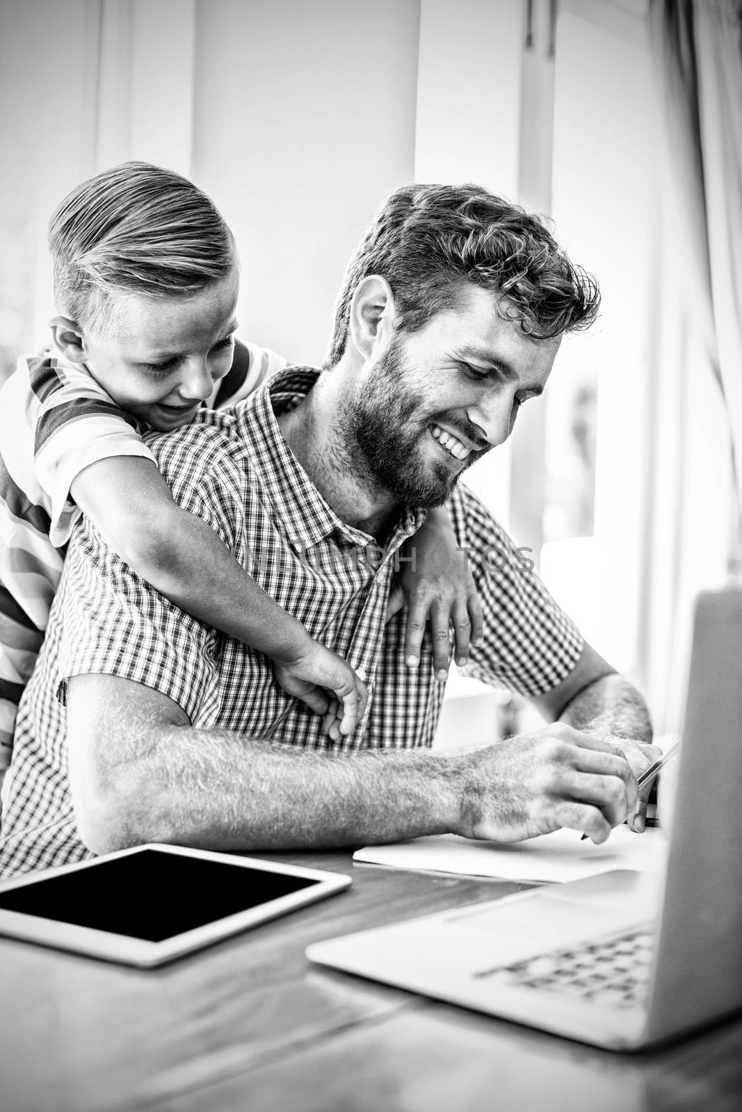 Smiling son leaning on father working with laptop and tablet at home 