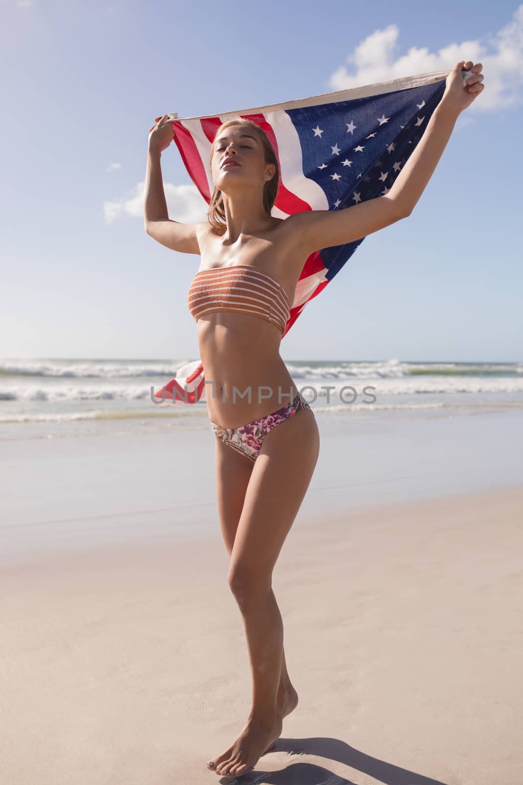 Front view of young woman holding waving American flag at beach on a sunny day