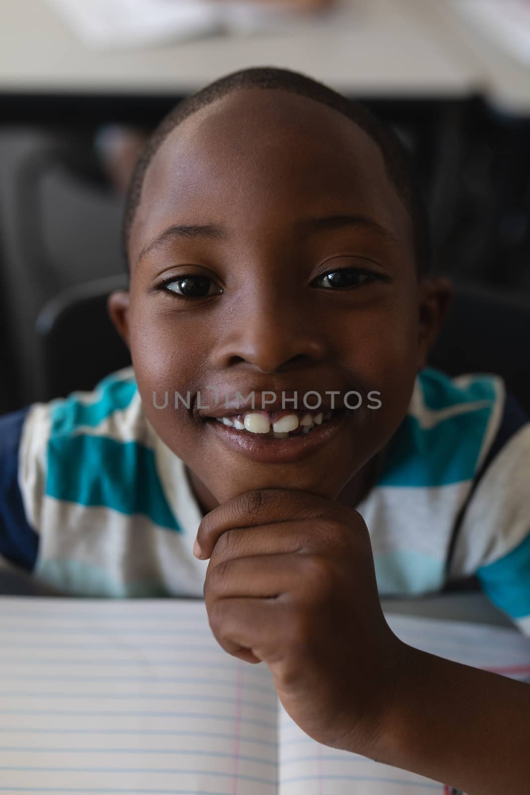 Close-up of black schoolboy with hand on chin sitting at desk and looking at camera in classroom of elementary school