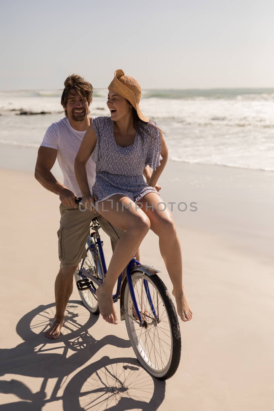 Front view of beautiful happy young woman sitting on man bicycle handlebars on beach in the sunshine