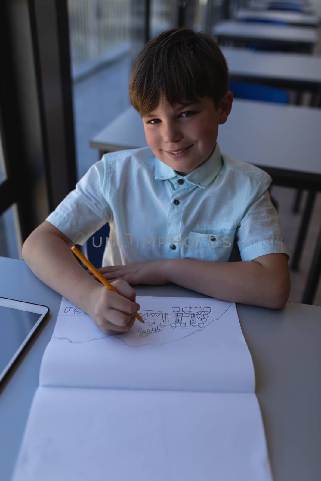 Front view of schoolboy drawing on notebook and looking at camera at desk in classroom of elementary school